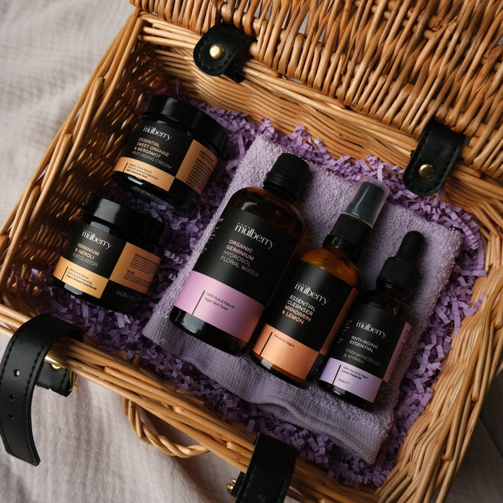 A wicker picnic basket lined with purple tissue paper and black leather straps, holding several skincare bottles and jars from the brand Meruity, arranged on a textured light gray fabric surface.